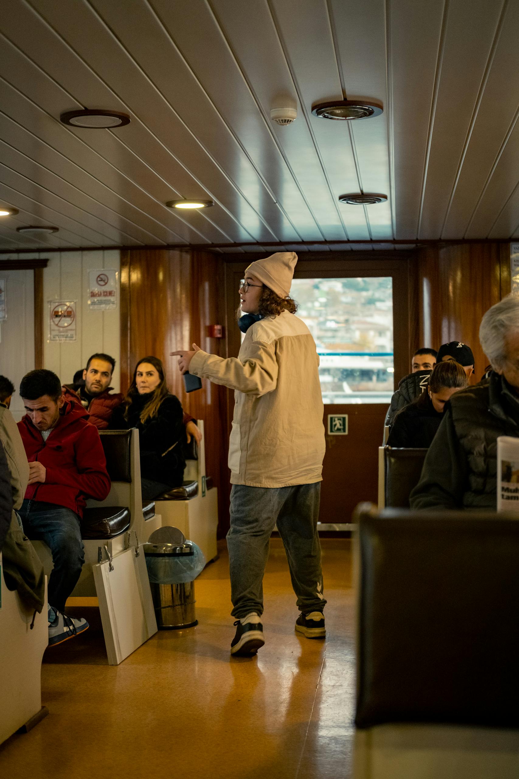 A relaxed scene inside a ferry with diverse passengers seated and a person standing.