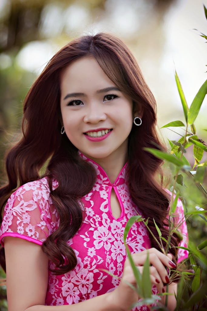 A smiling woman in a pink floral dress poses outdoors surrounded by greenery.