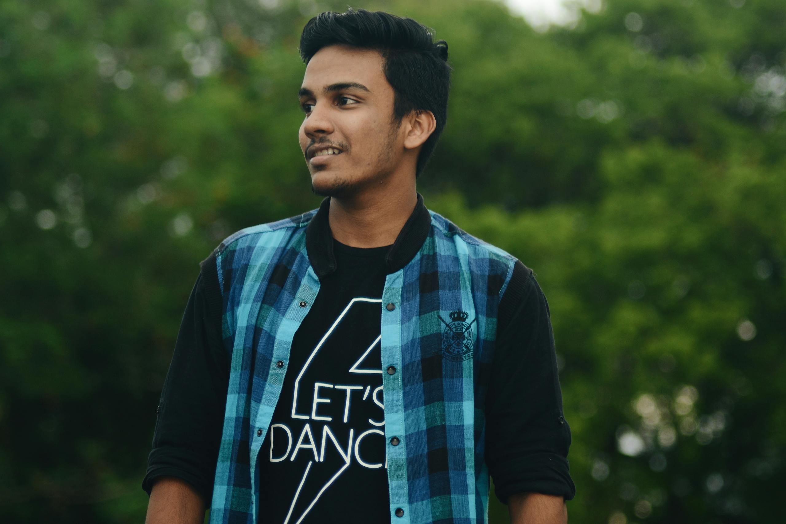 A young man in a casual plaid shirt smiling while standing outside against greenery.