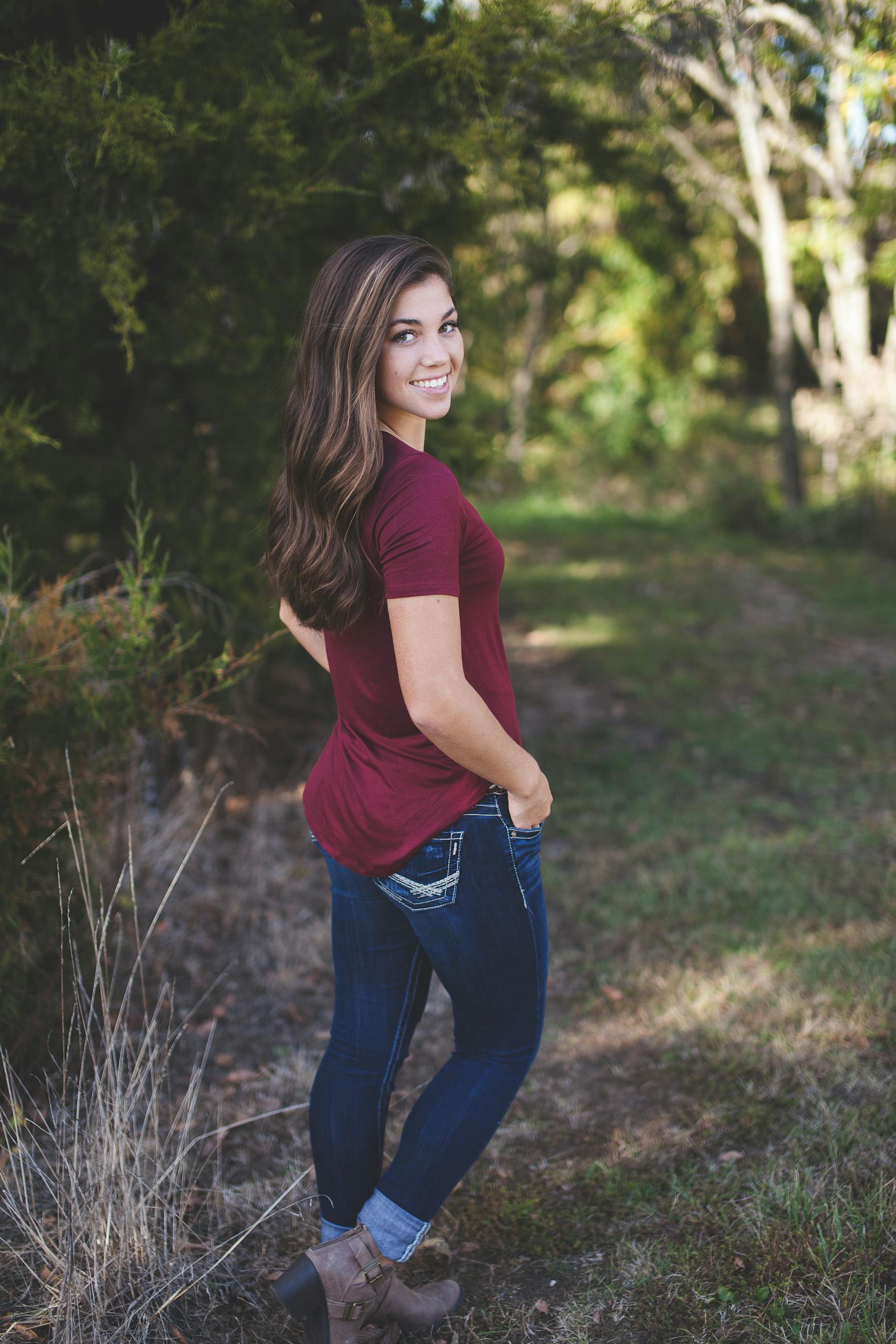 A young woman with long hair smiles outdoors in a forest setting, wearing casual attire.