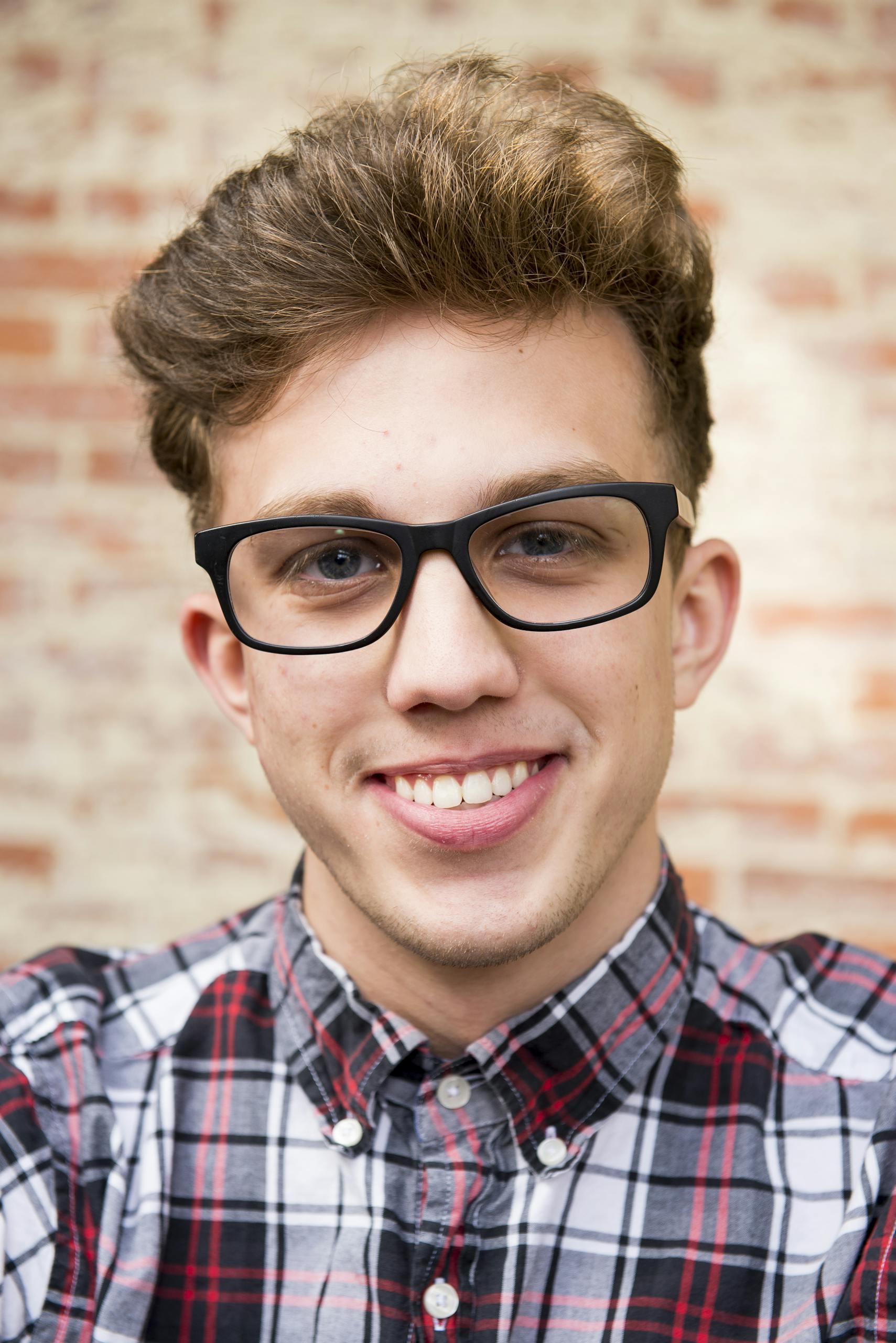 Portrait of a smiling young man wearing eyeglasses and plaid shirt against a brick wall background.